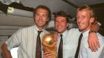 German team leader and head coach Franz Beckenbauer (L) stands with German midfielder and team captain Lothar Matthaeus (C) and German defender Andreas Brehme in the cabin of an airplane and present the World Cup trophy on their way to Germany, 9 July 1990. Germany had won the World Cup final against Argentina in Italy by a score of 1-0 and had won the world championship for the third time since 1954 and 1974. (Photo by Eilmes / DPA / dpa Picture-Alliance via AFP)