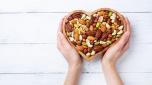 Womans hands holding heart shaped bowl with mixed nuts on white table top view. Healthy food and snack.