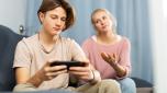 Interested excited teen boy playing online game on cell phone at home while upset mother trying to get his attention. Concept of family phubbing behavior
