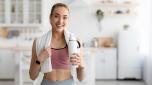 Covid-19, active sports at home and thirst, healthcare. Smiling young blonde woman in sportswear resting after workout in minimalist living room interior with towel on shoulders and bottle of water