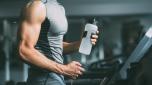 Unrecognisable young man in sportswear running on treadmill at gym and holding bottle of water
