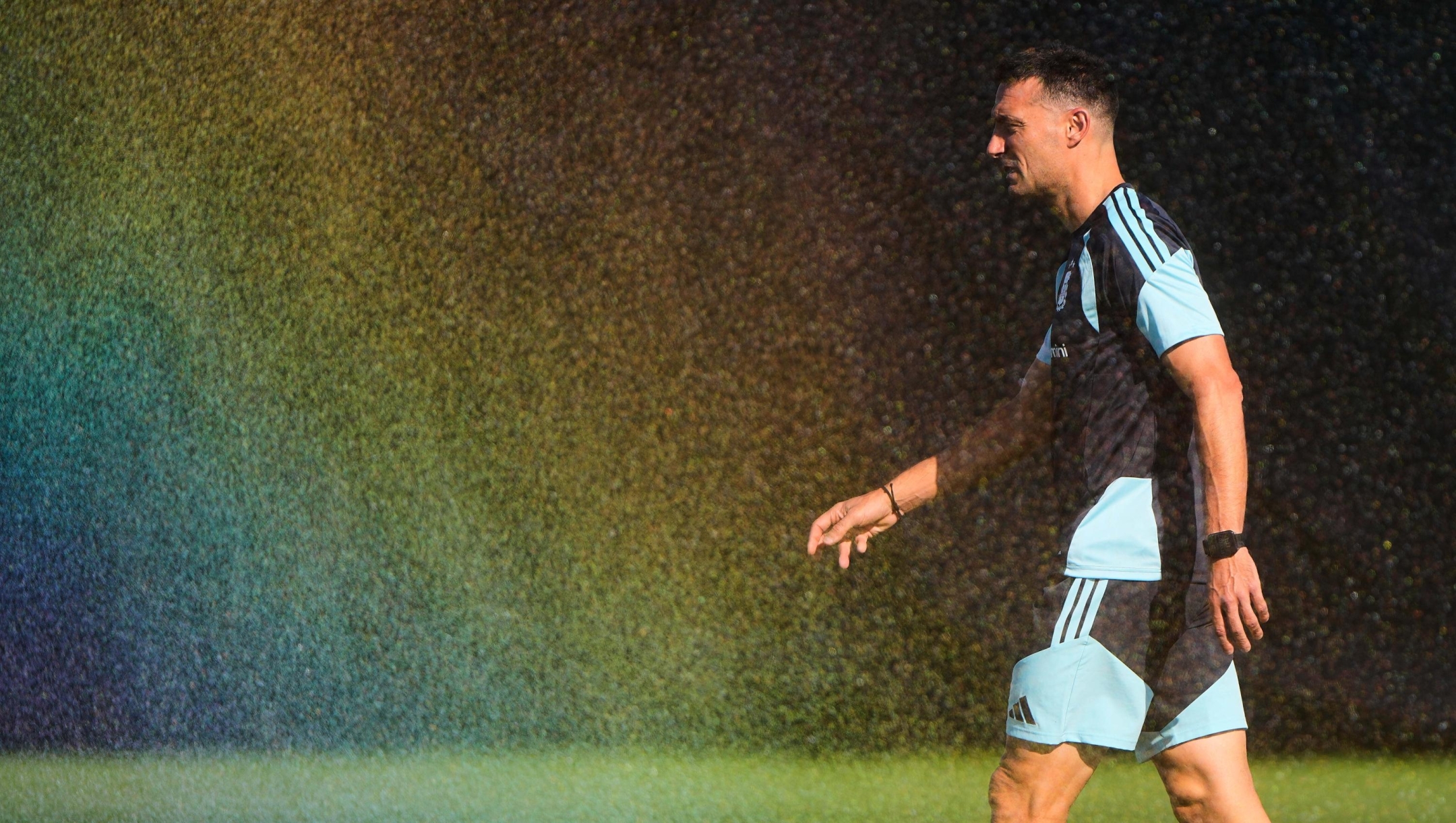 Water droplets from a sprinkler form a rainbow as Argentina head coach Lionel Scaloni walks across the pitch during training for the World Cup soccer tournament Wednesday, June 24, 2026, in Kansas City, Kan. (AP Photo/Charlie Riedel)