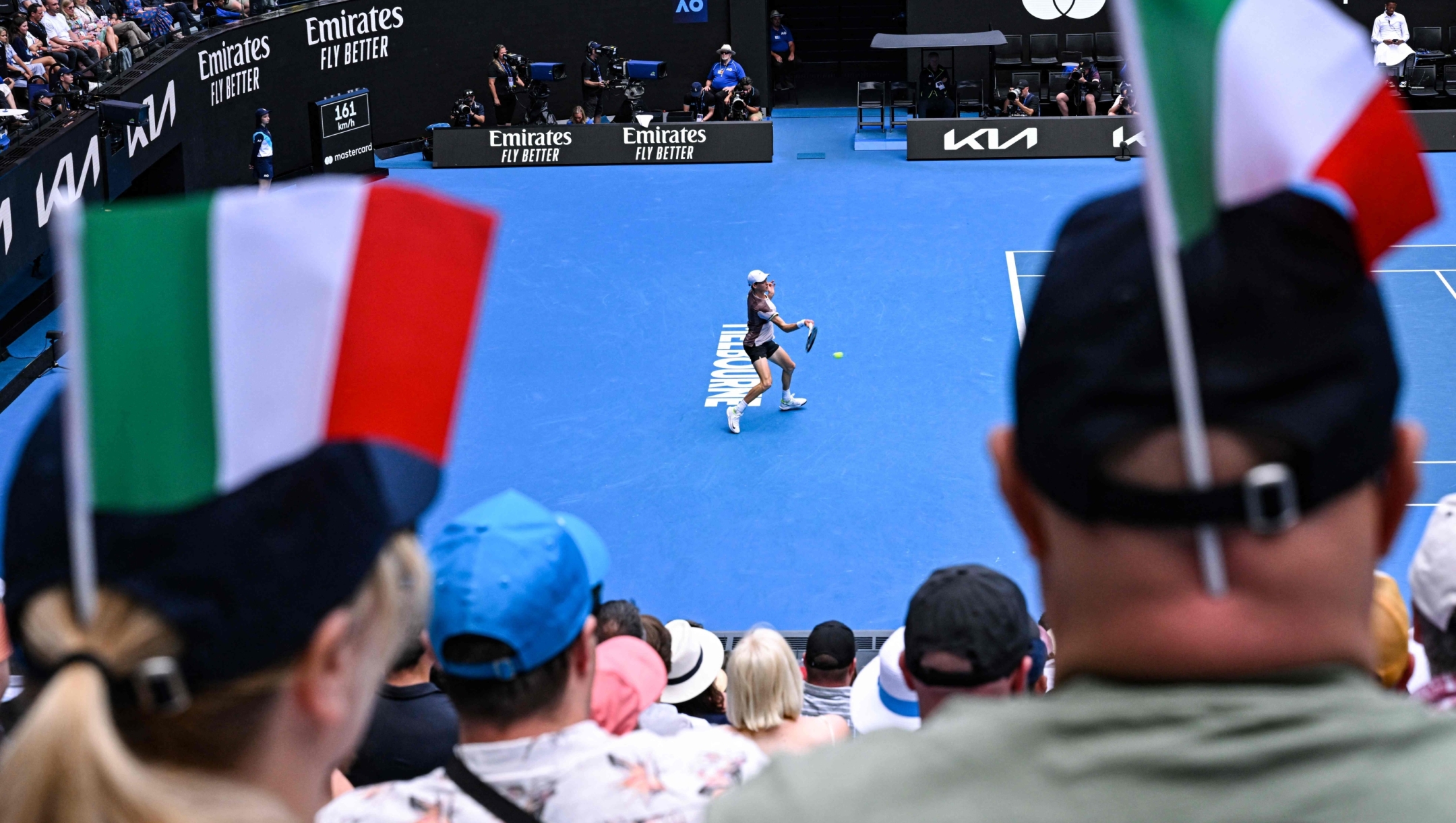 Spectators with Italian flags look at Italy's Jannik Sinner hitting a return to Serbia's Novak Djokovic during their men's singles semi-final match on day 13 of the Australian Open tennis tournament in Melbourne on January 26, 2024. (Photo by WILLIAM WEST / AFP) / -- IMAGE RESTRICTED TO EDITORIAL USE - STRICTLY NO COMMERCIAL USE --