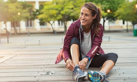 Mature fitness woman tie shoelaces on road. Cheerful runner sitting on floor on city streets with mobile and earphones wearing sport shoes. Active latin woman tying shoe lace before running.