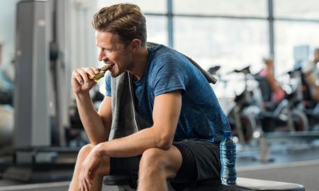 Sweaty young man eating energy bar at gym. Handsome mid guy enjoying chocolate after a heavy workout in fitness studio. Fit man biting a snack and resting on bench.