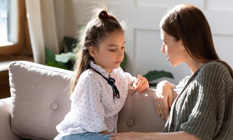 Upset small schoolgirl having trustful conversation with compassionate young mother, sitting together on sofa. Wise mommy comforting soothing little child daughter, overcoming problems at home.