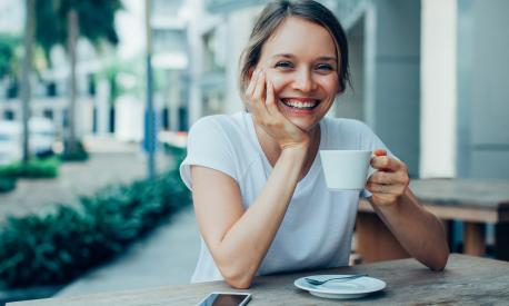 Closeup portrait of smiling young beautiful woman looking at camera, leaning head on hand and drinking coffee at cafe table outdoors with street view in background. Front view.