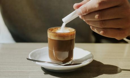 a young man ads some sugar to his machiatto sitting at the table of a sidewalk cafe, in a panoramic format to use as web banner or header