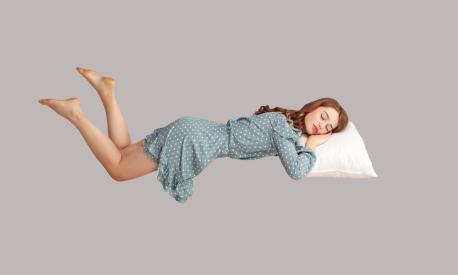 Relaxed girl in vintage ruffle dress levitating in mid-air, sleeping on stomach lying comfortable cozy on pillow, keeping eyes closed, watching peaceful dream. indoor studio shot isolated on gray