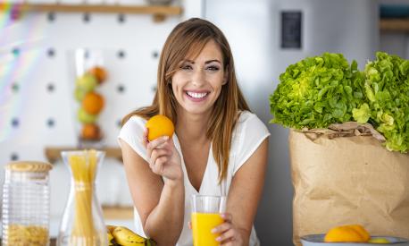 Young joyful woman drinking orange juice and standing near a kitchen table. Close up of a woman drinking juice in her kitchen. Fit smiling young woman preparing healthy fruit juice