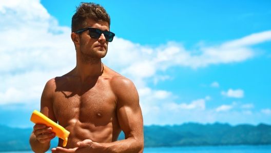 Man Skin Care In Summer. Handsome Male With Sexy Body In Sunglasses Applying UV Protective Sunscreen Lotion Before Sunbathing, Tanning At Beach Using Solar Sun Block Protection Cream For Healthy Tan.