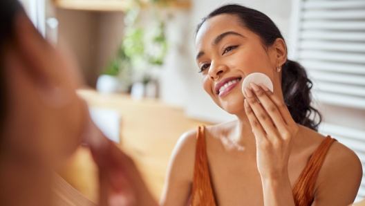 Portrait of latin young beauty woman looking at mirror while cleaning her face with cotton disc in bathroom. Beautiful multiethnic woman taking good care of her face while standing against mirror in bathroom. Enjoying facial skincare routine concept.