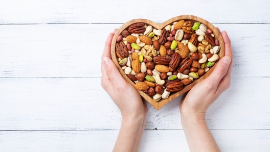 Womans hands holding heart shaped bowl with mixed nuts on white table top view. Healthy food and snack.