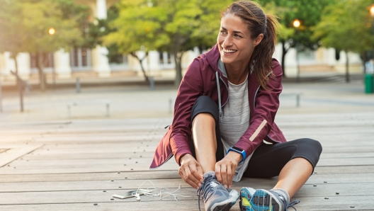 Mature fitness woman tie shoelaces on road. Cheerful runner sitting on floor on city streets with mobile and earphones wearing sport shoes. Active latin woman tying shoe lace before running.