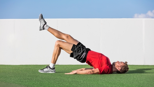 Fitness man doing bodyweight glute single leg floor bridge lift exercises. Fit athlete training glutes muscles with one-legged floor bridge butt raise in summer outdoor gym on grass.