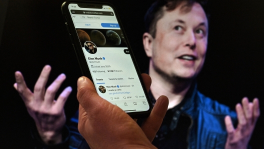 (FILES) In this file photo illustration, a phone screen displays the Twitter account of Elon Musk with a photo of him shown in the background, on April 14, 2022, in Washington, DC. - Twitter shares slid late on July 7, 2022 after a Washington Post report that Elon Musk's $44 billion deal to buy the social media giant is in danger. (Photo by Olivier DOULIERY / AFP)