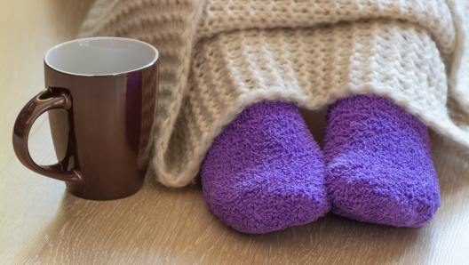 Cold concept with teacup near the legs which are covered with fluffy warm blanket and wearing fluffy warm purple socks