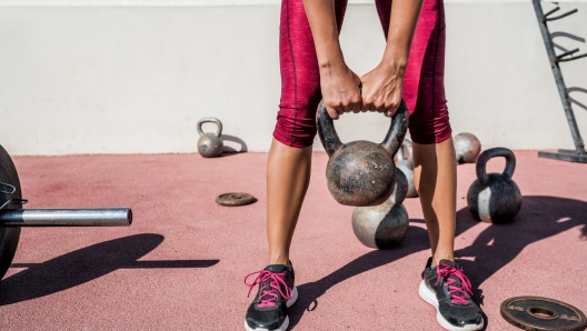 Woman weightlifting kettlebell weight at outdoor fitness gym. Unrecognizable female athlete strength training legs, glutes and back lifting free weights. Closeup of lower body.