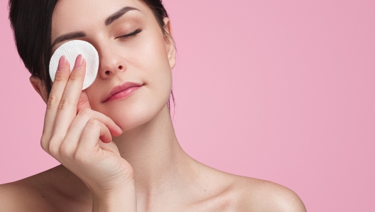 Crop attractive young female cleansing face with a cotton pad on a pink background in studio