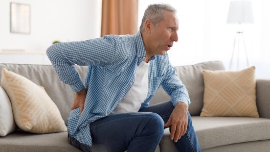 People, Healthcare and Treatment. Portrait of sick mature man suffering from acute side back pain, sitting on couch at home, person touching lower back aching area with hand. Health Problem Concept