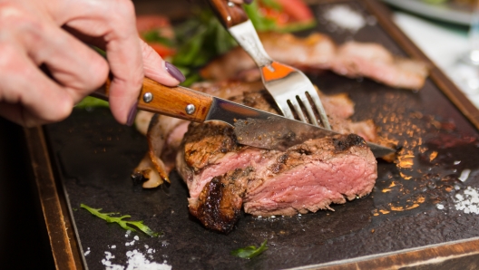 Woman hand holding knife and fork cutting grilled beef steak on stoned plate. Selective focus