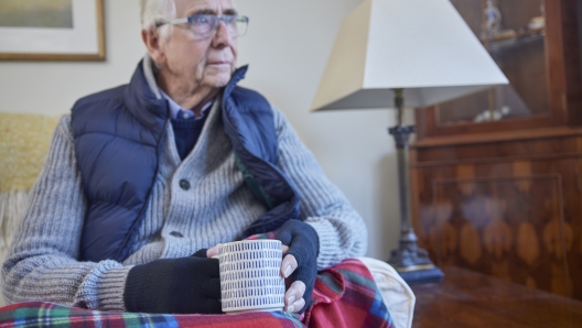 Senior Man Wearing Extra Clothes With Hot Drink Trying To Keep Warm At Home In Energy Crisis