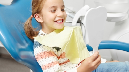 Little caucasian girl looks to the mirror and smiling after visiting of dentist. Happy child with a healthy teeth.