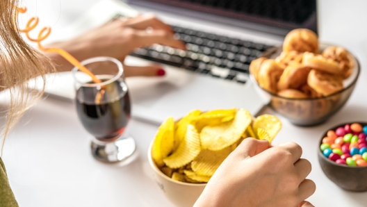 Girl works at a computer and eats fast food. Unhealthy food: chips, crackers, candy, waffles, cola. Junk food, concept.