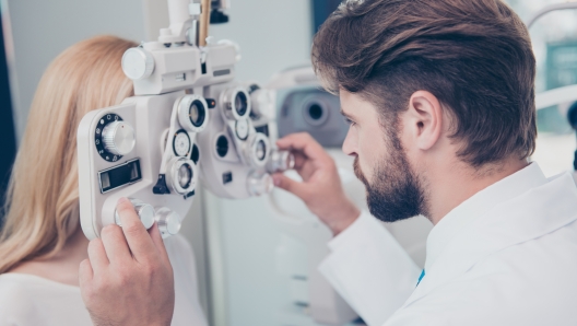 Close-up of brunet bearded doc optometrist adjusting phoropter for young blond female patient. Eye sight and healthcare