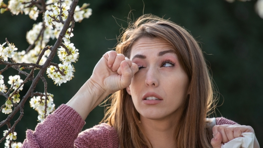 Young pretty girl rubbing eyes beside blooming tree in spring time. Itchy eyes from allergy