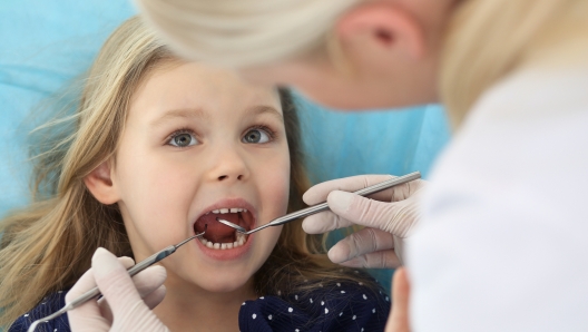 Little baby girl sitting at dental chair with open mouth and feeling fear during oral check up while doctor. Visiting dentist office. Medicine concept.