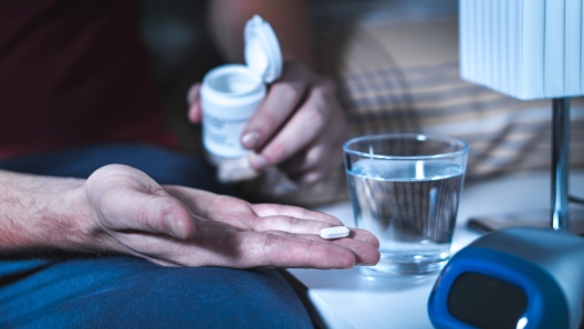 Man sitting on bed about to take sleeping pill or night medicine. Suffering from insomnia. Holding tablet and bottle in hand. Glass of water on nightstand in bedroom.