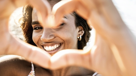 Close up image of smiling woman in swimwear on the beach making a heart shape with hands - Pretty joyful hispanic woman laughing at camera outside - Healthy lifestyle, self love and body care concept