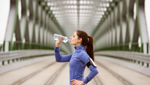 Beautiful young woman with smart phone and earphones, listening music, running in the city on green steel bridge, resting, drinking water from bottle.