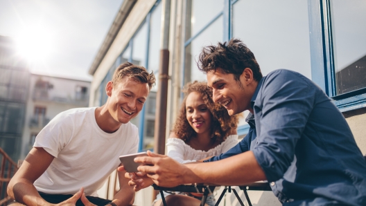 Three young friends sitting outdoors and looking at mobile phone. Group of people sitting at outdoor cafe and watching video on the smartphone.