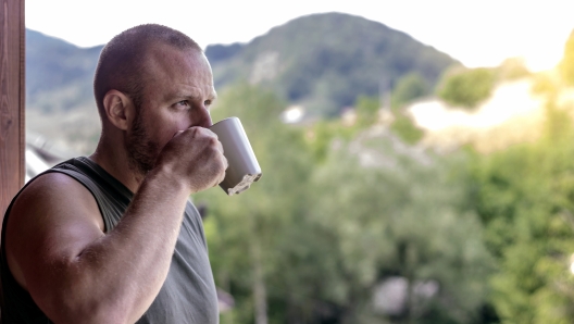 Photo of smiling young man standing on the balcony with cup of coffee.Handsome man having his morning coffee on a balcony and daydreaming.Caucasian man looking away and sipping coffee in white cup.