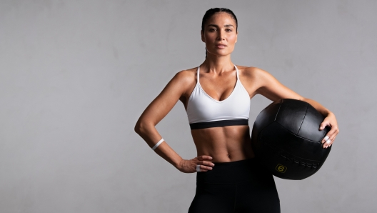 Portrait of beautiful mid adult woman looking at camera while holding heavy medicine ball isolated on grey background. Proud and fit woman standing on gray wall ready for gym exercise while looking at camera. Strong mixed race girl holding sports ball and relaxing after cross training workout with copy space.