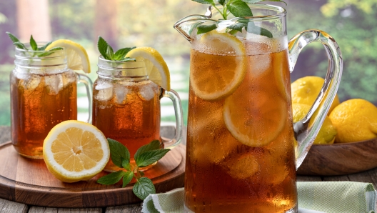 Pitcher of cold iced tea with mint, lemon slices and ice with two glasses ot tea on a wooden table and rural summer background