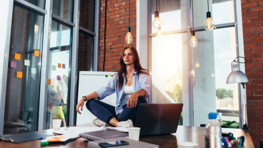 Young businesswoman meditating sitting on working table in lotus pose in her office.