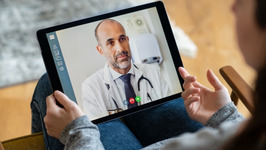 Back view of young woman making video call with her doctor while staying at home. Close up of patient sitting on armchair video conferencing with general practitioner on digital tablet. Sick girl in online consultation with a mature physician.