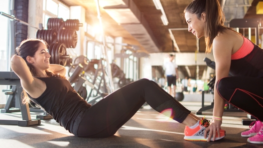 Young woman exercising sit-ups with assistance of female friend in gym.