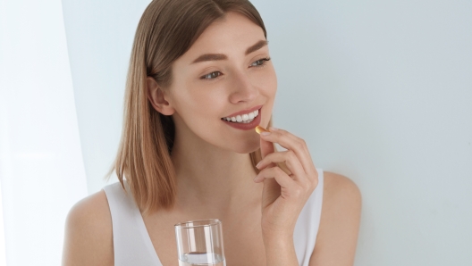 Woman taking vitamin pill with glass of fresh water indoors. Smiling girl taking omega 3 fish oil capsule, vitamin supplement. Diet nutrition concept