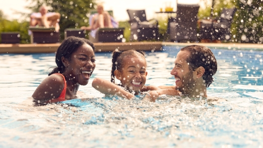 Smiling Mixed Race Family On Summer Holiday Having Fun Splashing In Outdoor Swimming Pool