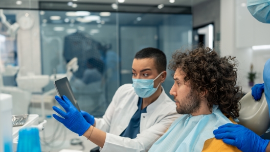 Focused handsome man with curly hair sitting on dentist's chair watching digital presentation on digital tablet device with two female dental professionals, dentist and her colleague at modern dental clinic.