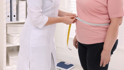 Nutritionist measuring overweight woman's waist with tape in clinic, closeup