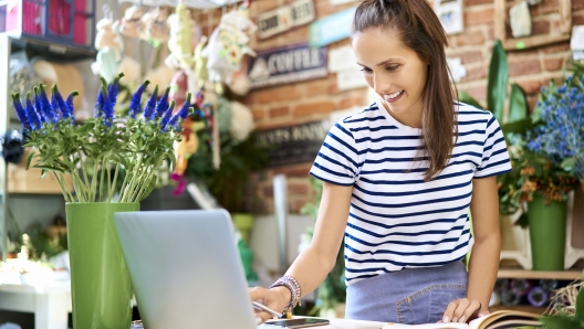 Cheerful florist looking at laptop and writing down client orders in notebook