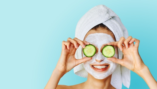 Beautiful young woman with facial mask on her face holding slices of fresh cucumber. Skin care and treatment, spa, natural beauty and cosmetology concept, over blue background with copy space