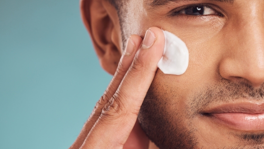 Closeup of one young indian man applying moisturiser lotion to his face while grooming against a blue studio background. Handsome guy using sunscreen with spf for uv protection. Rubbing facial cream on cheek for healthy complexion and clear skin