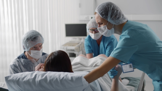 Woman giving birth with husband holds her hand in support and obstetricians assisting. Back view of medical staff in protective uniform helping pregnant woman in labor