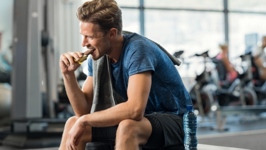 Sweaty young man eating energy bar at gym. Handsome mid guy enjoying chocolate after a heavy workout in fitness studio. Fit man biting a snack and resting on bench.